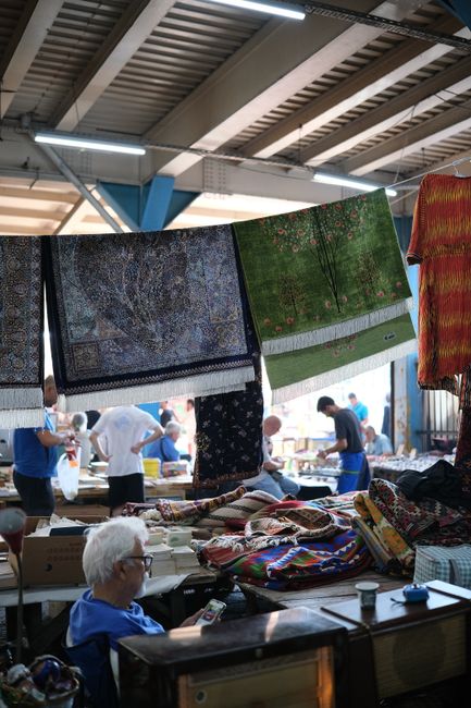 A person dressed in a blue denim jacket and matching cap is seated on a stool at an indoor market stall, surrounded by various items and textiles. The area is brightly lit with natural and artificial